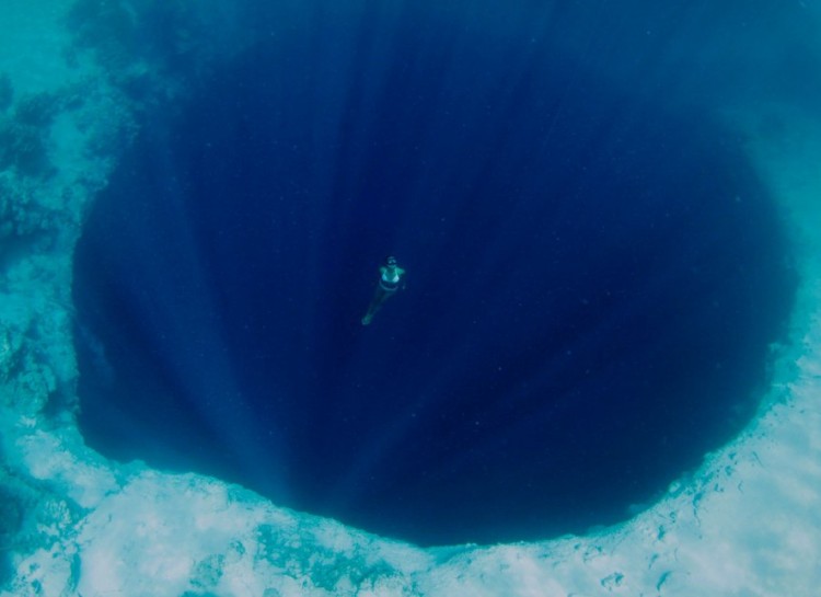 A lone diver floats above a massive underwater sinkhole, known as a blue hole.